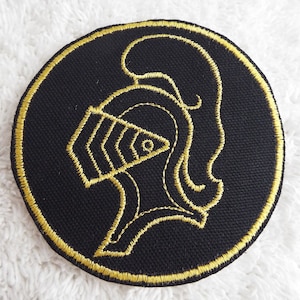 May include: A black circular patch with a gold embroidered design of a knight's helmet.