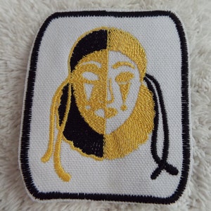 May include: A white embroidered patch featuring a black and gold face mask design. The mask is half black and half gold, with a white outline. The mask has a black and gold braid on each side.
