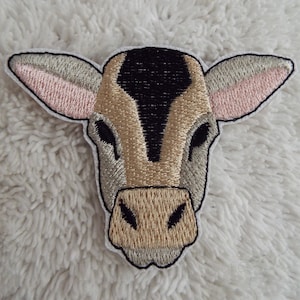May include: Embroidered patch of a brown and black cow's face with pink ears. The patch is on a white fluffy surface.