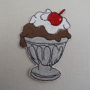 Puede incluir: Parche bordado para planchar de un helado de vainilla con salsa de chocolate y una cereza roja en la parte superior en un plato plateado.