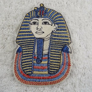 Puede incluir: Parche bordado para planchar de la cabeza de un faraón egipcio con un tocado azul y dorado. La cara del faraón se representa de perfil con una expresión seria.