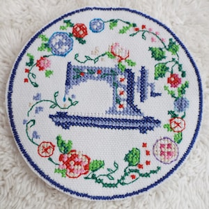 May include: A white circular embroidery hoop with a blue sewing machine design surrounded by colorful flowers and vines.