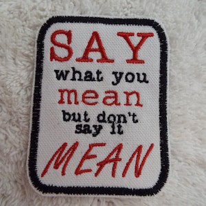 May include: White fabric patch with black stitching around the edges. The patch has red embroidered text that reads "SAY what you mean but don't say it MEAN".