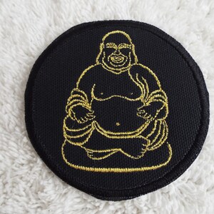 May include: A black fabric patch with a gold embroidered outline of a smiling Buddha figure.