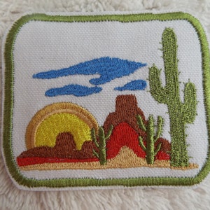May include: Embroidered patch with a desert scene featuring a yellow sun, brown mountains, a red rock formation, green cacti, and blue clouds against a white background. The patch is surrounded by a green border.