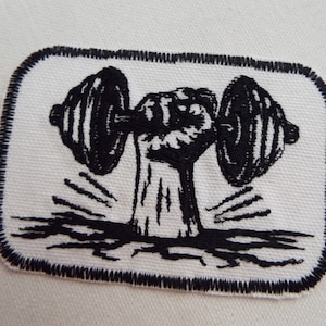 May include: Black and white embroidered patch of a fist lifting a barbell. The patch has a black border and is on a white background.