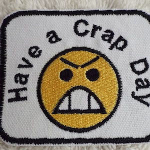 Może przedstawiać: Biała naszywka haftowana z czarną obwódką. Naszywka przedstawia żółtą złą twarz z czarnymi oczami i czarnymi ustami. Tekst "Have a Crap Day" jest wyszyty wokół twarzy.