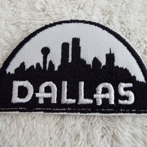 May include: White and black embroidered patch with a skyline of Dallas, Texas. The text "DALLAS" is in white lettering.