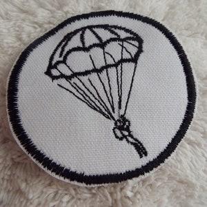 May include: A white round patch with a black embroidered image of a parachute. The parachute is open and has a person hanging from it.