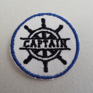 May include: A circular patch with a white background and a blue stitched border. The patch features a black ship's wheel design with the word "CAPTAIN" embroidered in black across the center.