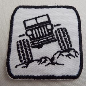May include: Square white patch with a black embroidered design of a Jeep off-roading. The Jeep is angled, with large tyres and a detailed front grill. The patch has a black stitched border.