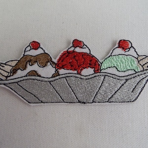 Puede incluir: Un parche bordado de un banana split con tres bolas de helado: chocolate, fresa y menta. El helado está en un plato plateado con bordes ondulados.