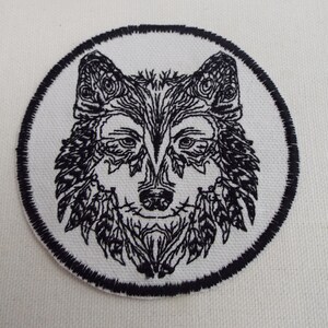 以下が含まれることがあります： 複雑なディテールで刺繍された黒と白の狼の顔のパッチ。狼の顔は黒い円に囲まれています。