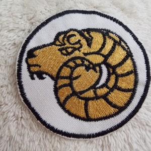 Op de afbeelding: Een gouden geborduurde patch met een zwarte omtrek, die een rammenkop met gekrulde hoorns voorstelt. De patch is rond en heeft een witte achtergrond.