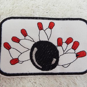 Puede incluir: Un parche bordado blanco con una bola de bolos negra y diez bolos blancos con puntas rojas.