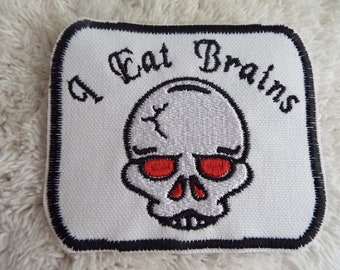 May include: White embroidered patch with a black border. The patch features a skull with red eyes and the text "I Eat Brains" in black.