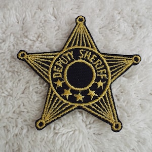 May include: A gold embroidered star-shaped patch with the words "DEPUTY SHERIFF" around a black circle. The patch is on a white fuzzy surface.