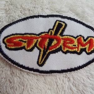 May include: Oval-shaped embroidered patch with the word "STPEM" in red and gold lettering, outlined in black. The patch is on a white background with a black border.