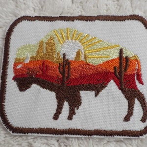 May include: Embroidered patch featuring a brown silhouette of a buffalo standing in front of a sunset landscape with a yellow sun and red, orange, and yellow hills. The patch has a brown border.