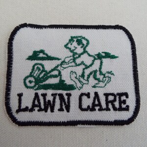 Könnte beinhalten: Weißer gestickter Aufnäher mit einer grünen Cartoon-Figur, die einen Rasen mäht. Der Text "LAWN CARE" ist in Schwarz unter dem Bild gestickt.