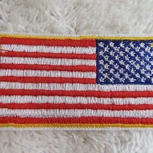 Può includere: Un piccolo patch ricamato della bandiera americana con strisce rosse e bianche e un campo blu di stelle. Il patch ha un bordo giallo.