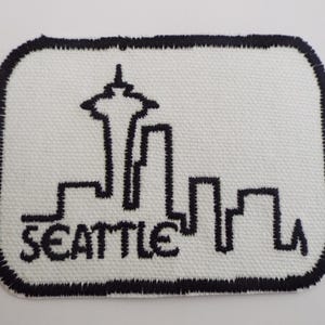 May include: White rectangular patch with a black embroidered border. The patch features a black outline of the Seattle skyline, including the Space Needle, with the city's name embroidered below.
