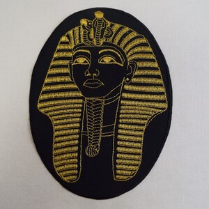 May include: A gold embroidered patch of an Egyptian pharaoh's head with a black background.
