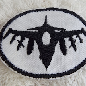 May include: A white oval patch with a black embroidered silhouette of a fighter jet. The jet is facing forward with its wings spread out.