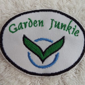 May include: White oval embroidered patch with black edging. The patch says "Garden Junkie" in green cursive lettering. A green stylized leaf design with a blue circle is in the centre.
