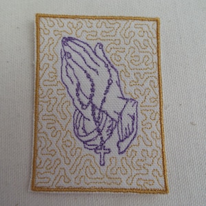 May include: Purple embroidered patch of praying hands with a rosary and cross on a white background with a gold embroidered border.