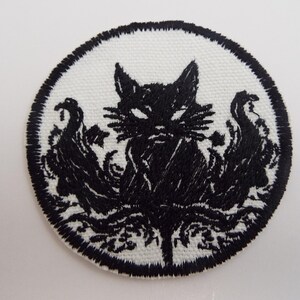 May include: A circular embroidered patch featuring a black cat silhouette centered on a white background. The cat is surrounded by intricate black swirling patterns and foliage, all enclosed within a thick, stitched black circular border.