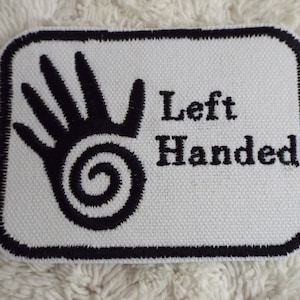 Pode incluir: Etiqueta branca bordada com contorno preto. A etiqueta apresenta uma mão preta estilizada com um desenho em espiral e o texto "Left Handed".
