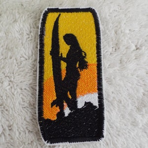 May include: Black and white embroidered patch of a person standing on a beach holding a surfboard. The background is a yellow and orange sunset.