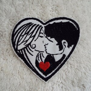 May include: Embroidered patch in the shape of a heart with a black and white illustration of a couple kissing. The couple is surrounded by a red heart.