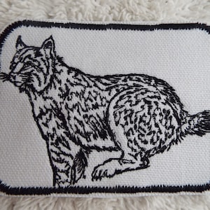 May include: Black and white embroidered patch of a lynx with a white background and black border.