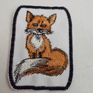 May include: Embroidered patch featuring a fox with a white chest and a bushy tail. The fox is sitting on a white background with a black border.