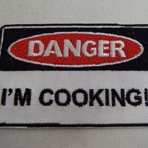 Pode incluir: Etiqueta branca bordada com borda preta e oval vermelho com texto branco "DANGER" dentro. A parte inferior da etiqueta diz "I'M COOKING!"