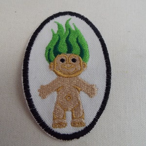 Puede incluir: Un parche bordado con un troll de pelo verde y contorno negro. El troll sonríe y tiene un cuerpo marrón claro.