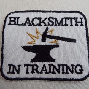 Op de afbeelding: Geborduurde patch met een witte achtergrond en een zwarte rand. De patch heeft een zwart aambeeld- en hamerontwerp met goudkleurige vonken en de woorden "BLACKSMITH IN TRAINING" in het zwart.