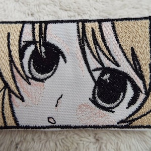 May include: Embroidered patch featuring a close-up of an anime character's face with large eyes. The character has blonde hair and is looking directly at the viewer.