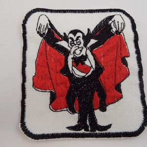 May include: A square embroidered patch featuring a cartoon vampire character with black hair, a black suit, and a red-lined cape. The character is depicted with arms raised, displaying the interior of the cape against a white fabric background.