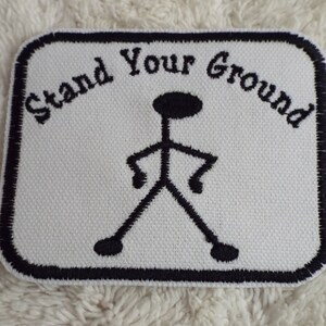 May include: White embroidered patch with black stitching. The patch features a stick figure with the text "Stand Your Ground" around the figure.