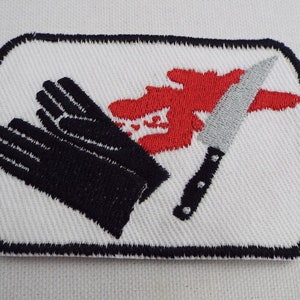 May include: A white embroidered patch with a black knife, black gloves, and a red blood splatter.