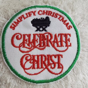 May include: Round white embroidered patch with green and red detailing. The patch reads "SIMPLIFY CHRISTMAS" and "CELEBRATE CHRIST" in red, with a black nativity scene illustration. It has a green border.