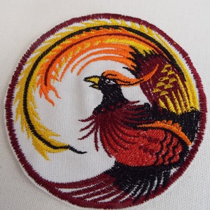May include: Embroidered patch featuring a stylized phoenix with red, orange, and yellow feathers, surrounded by a maroon circle.