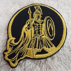 Könnte beinhalten: Ein gold gestickter Aufnäher mit einem Krieger, der einen Speer und einen Schild hält, auf schwarzem Hintergrund.
