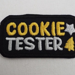 Może przedstawiać: Czarna łatka z tkaniny z białym i żółtym haftem. Łatka zawiera napis "Cookie Tester" z białą gwiazdą i żółtym drzewem sosnowym.