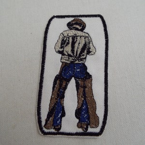 May include: Embroidered patch depicting a person wearing a brown cowboy hat, a light brown jacket, blue jeans, and brown chaps. The patch is rectangular with a black border.