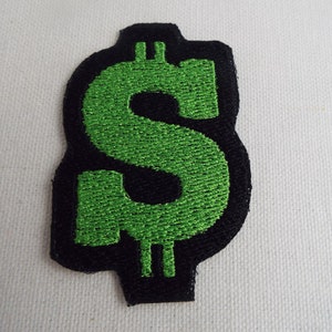 Puede incluir: Parche bordado con signo de dólar verde con contorno negro. El parche está hecho de tela y tiene un borde cosido.
