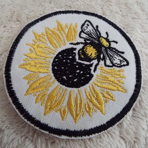 May include: A round embroidered patch with a black outline. The center of the patch features a yellow sunflower with a black center. A black and yellow bee is perched on the sunflower.